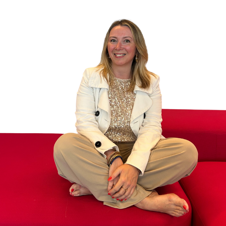 Femme souriante assise en tailleur sur un canapé rouge, portant un pull beige et une veste blanche.